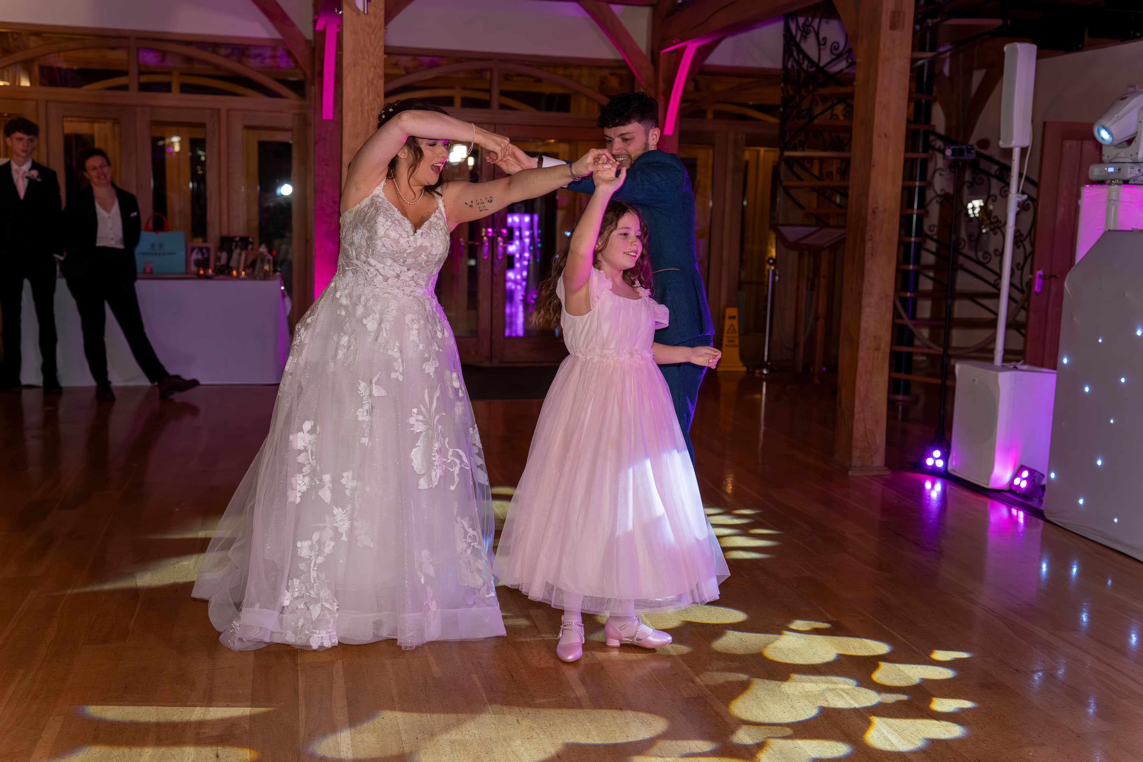 A bride and groom dance with a young guest on a wooden reception floor.